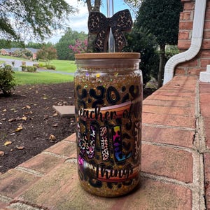 May include: A clear glass tumbler with a bamboo lid and a clear straw. The tumbler is decorated with gold glitter and the text "Teacher of all things" with colorful boot designs. A wooden bow-shaped charm is attached to the straw.