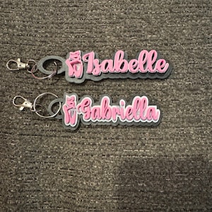 May include: Two pink and gray keychains with a ballerina shoe charm. The keychains are personalized with the names "Isabelle" and "Gabriella".