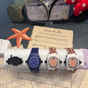 May include: A display of Disney-themed accessories, including two pairs of character ears and three MagicBand+ wristbands. The wristbands feature Mickey Mouse designs and leopard print patterns. A starfish and sheet music are also visible.