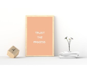 May include: A framed print with the words "TRUST THE PROCESS" in white on a peach background. A wooden clock and a small vase of flowers sit nearby on a white surface. The frame is light brown.