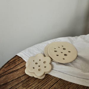 May include: A collection of beige ceramic coasters, including a flower-shaped stack and two round ones with holes. They rest on a white cloth atop a wooden table. The coasters are a neutral colour.