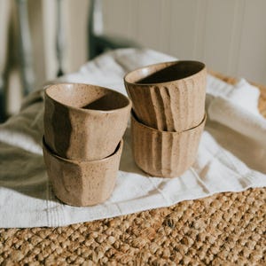 May include: Four small, beige ceramic bowls with a speckled finish are stacked on a white cloth. Two bowls have a ribbed texture, while the other two have a faceted design. The bowls are part of a kitchenware set.