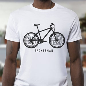 K&ouml;nnte beinhalten: Wei&szlig;es T-Shirt mit einer schwarzen Silhouette eines Fahrrads und dem Text "Spokesman" auf der Vorderseite.
