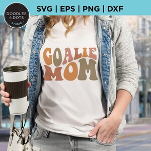 K&ouml;nnte beinhalten: Ein wei&szlig;es T-Shirt mit dem Text "Goalie Mom" in bunten Retro-Lettern. Die Buchstaben sind in Braun-, Orange- und Gelbt&ouml;nen gehalten.