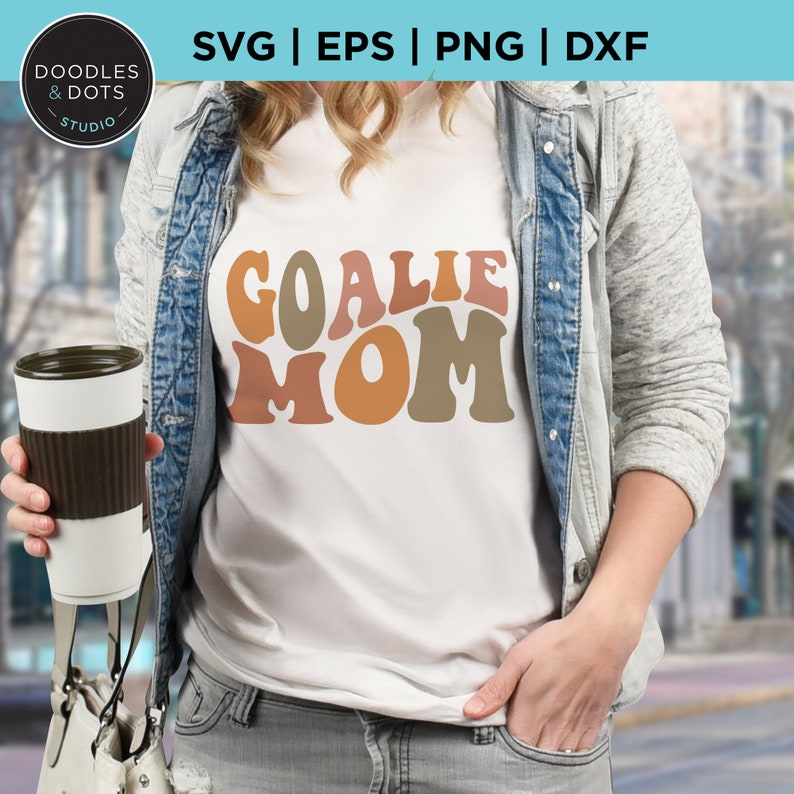K&ouml;nnte beinhalten: Ein wei&szlig;es T-Shirt mit dem Text "Goalie Mom" in bunten Retro-Lettern. Die Buchstaben sind in Braun-, Orange- und Gelbt&ouml;nen gehalten.