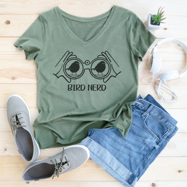 May include: A green v-neck t-shirt with a black graphic of binoculars with two birds inside. The text "BIRD NERD" is printed below the binoculars. The shirt is laying on a wooden surface with a pair of blue jeans, a pair of sneakers, and a potted plant.