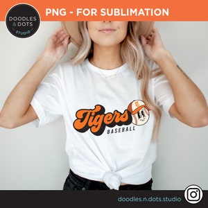 K&ouml;nnte beinhalten: Wei&szlig;es T-Shirt mit einer Retro-Baseballgrafik in Orange und Schwarz, auf der "Tigers Baseball" steht, sowie einem Cartoon-Baseball mit einem Gesicht, das eine Baseballkappe tr&auml;gt.