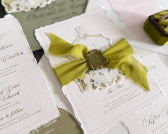 May include: A collection of wedding stationery featuring white textured paper with deckled edges, olive green envelopes, and a central invitation tied with a frayed olive green silk ribbon and a gold wax seal, displaying printed text and floral motifs.
