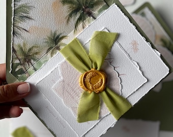 May include: Wedding invitation suite featuring a green envelope with a palm tree print. The invitation is layered with textured white paper, a green ribbon, and a gold wax seal. The suite includes additional cards.