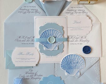 May include: A wedding invitation suite with a seashell theme. The invitation features a blue and white seashell with a blue wax seal. The RSVP card has a blue wax seal and the text "Please Reply". The envelope is light blue with a white wax seal and the text "Emily van deer Woodsen".