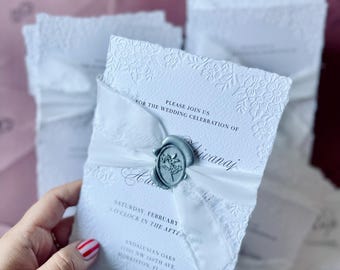 May include: White wedding invitation with embossed floral design, tied with a white satin ribbon and sealed with a silver wax seal. The invitation includes the text "PLEASE JOIN US FOR THE WEDDING CELEBRATION OF" and event details.