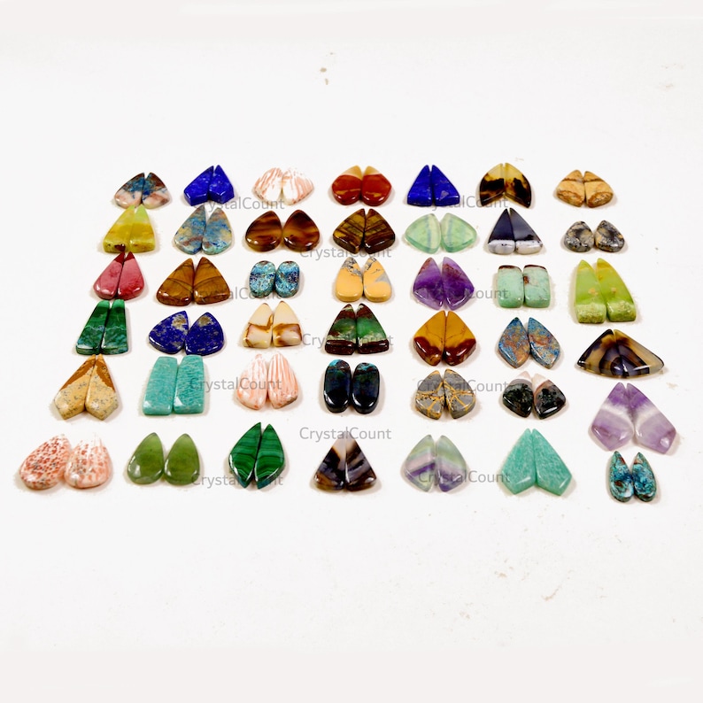 May include: A collection of numerous pairs of teardrop-shaped polished gemstones arranged in a grid. The stones feature a wide variety of colors, including blue, green, purple, brown, yellow, and pink, with various natural patterns and textures.
