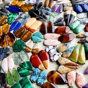 May include: A collection of assorted gemstone pendants in various shapes and colors, including tiger's eye, malachite, lapis lazuli, amethyst, and jasper. Each piece features a small drilled hole for stringing, showcasing polished surfaces.