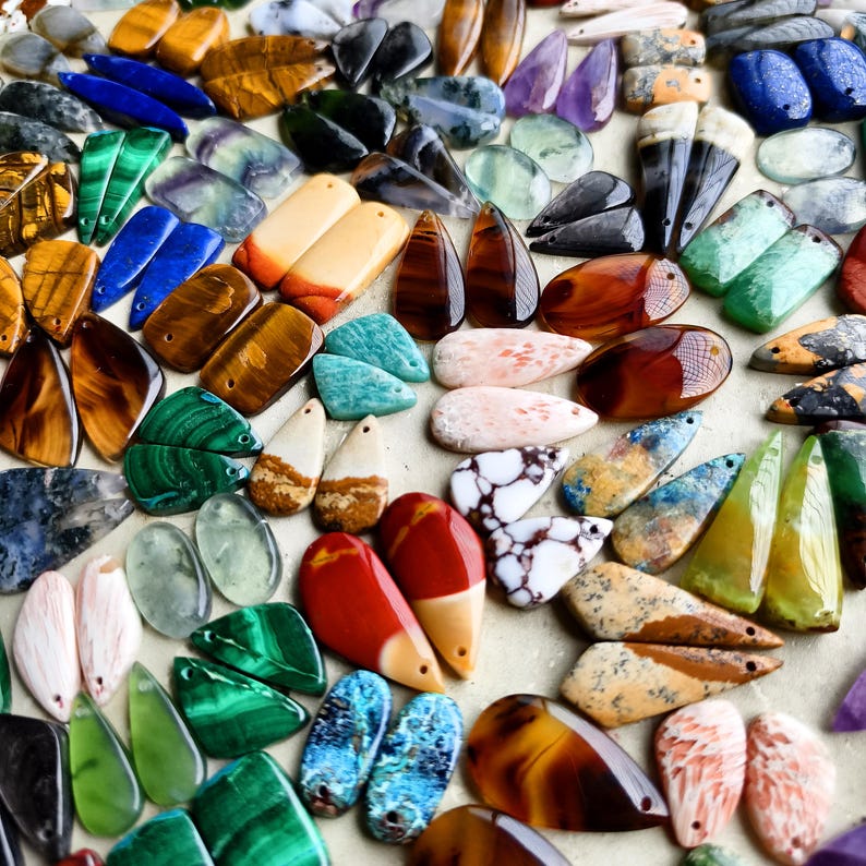 May include: A collection of assorted gemstone pendants in various shapes and colors, including tiger's eye, malachite, lapis lazuli, amethyst, and jasper. Each piece features a small drilled hole for stringing, showcasing polished surfaces.