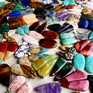 May include: A large collection of polished gemstone pendants in various shapes and colors, including tiger eye, malachite, amethyst, turquoise, and jasper. Each piece features a small drilled hole for stringing, showcasing natural stone patterns.