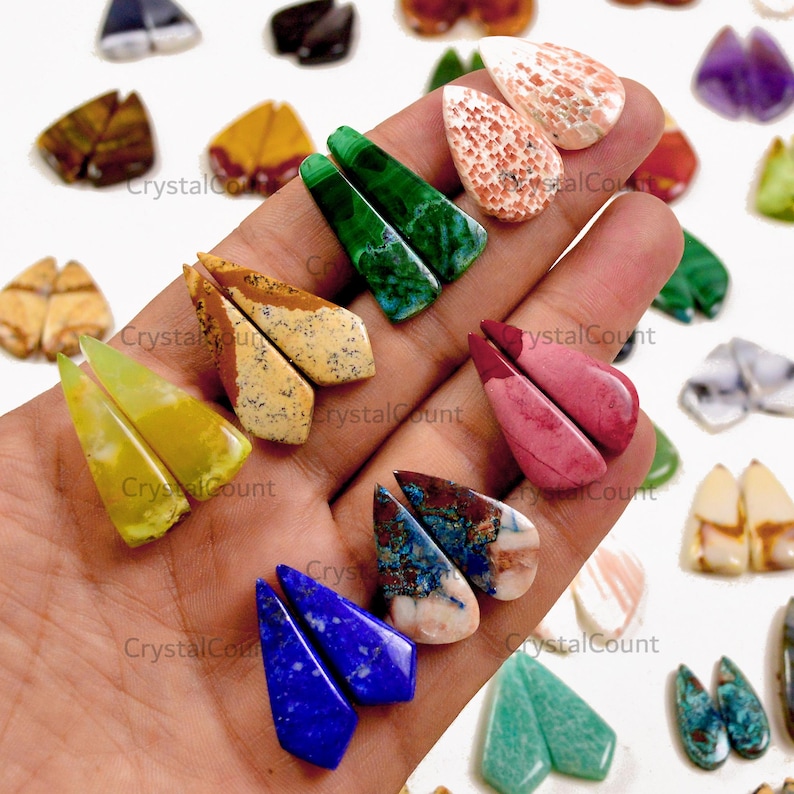 May include: A collection of polished, teardrop-shaped gemstone cabochons in various colors including green, yellow, pink, blue, and brown. The stones are arranged in pairs, featuring unique natural patterns, textures, and mineral inclusions.