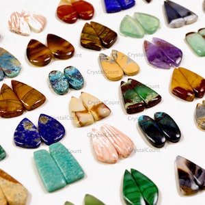 May include: A collection of assorted teardrop-shaped gemstone cabochons in various colors and patterns, including blue lapis lazuli, green malachite, brown tiger eye, purple amethyst, and striped agate, arranged in pairs on a white surface.