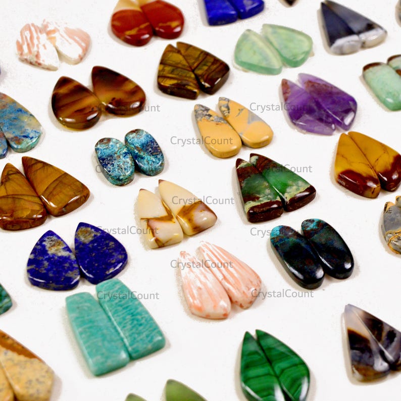 May include: A collection of assorted teardrop-shaped gemstone cabochons in various colors and patterns, including blue lapis lazuli, green malachite, brown tiger eye, purple amethyst, and striped agate, arranged in pairs on a white surface.