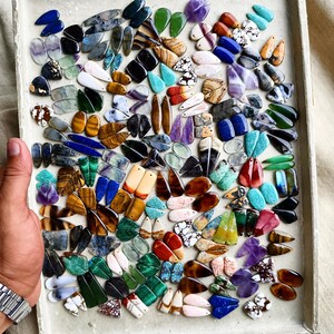 May include: A large collection of assorted gemstone pendants in various shapes, colors, and patterns, including teardrop, oval, and rectangular cuts. The stones feature diverse textures and mineral inclusions, arranged in a dense grid on a tray.