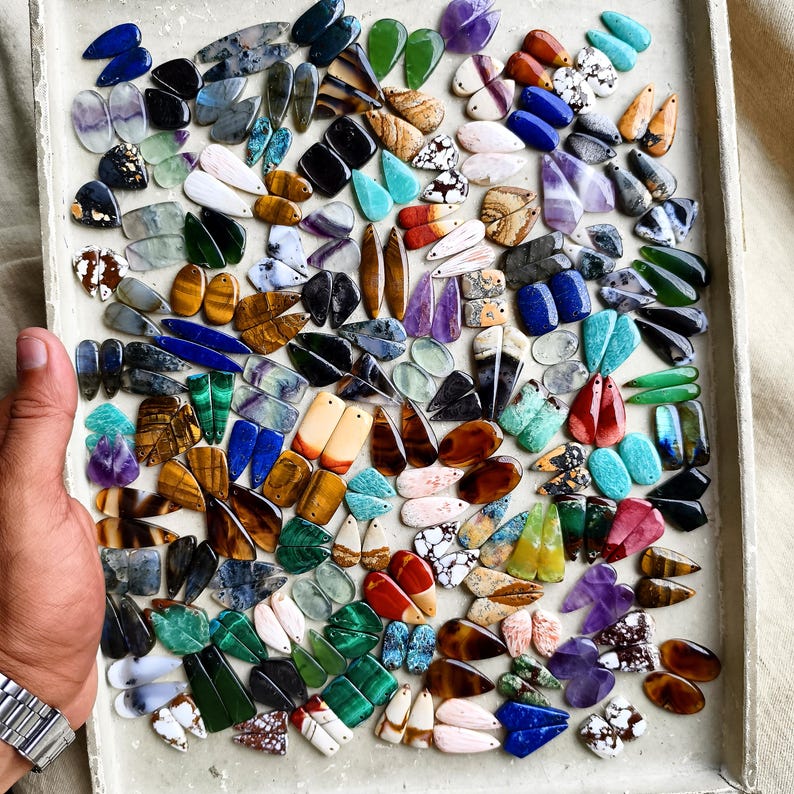 May include: A large collection of assorted gemstone pendants in various shapes, colors, and patterns, including teardrop, oval, and rectangular cuts. The stones feature diverse textures and mineral inclusions, arranged in a dense grid on a tray.