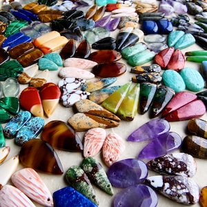 May include: A large collection of polished gemstone pendants in various shapes and colors, including teardrop, oval, and rectangular forms. The stones feature diverse patterns and hues such as deep purple, bright blue, green, brown, and white.