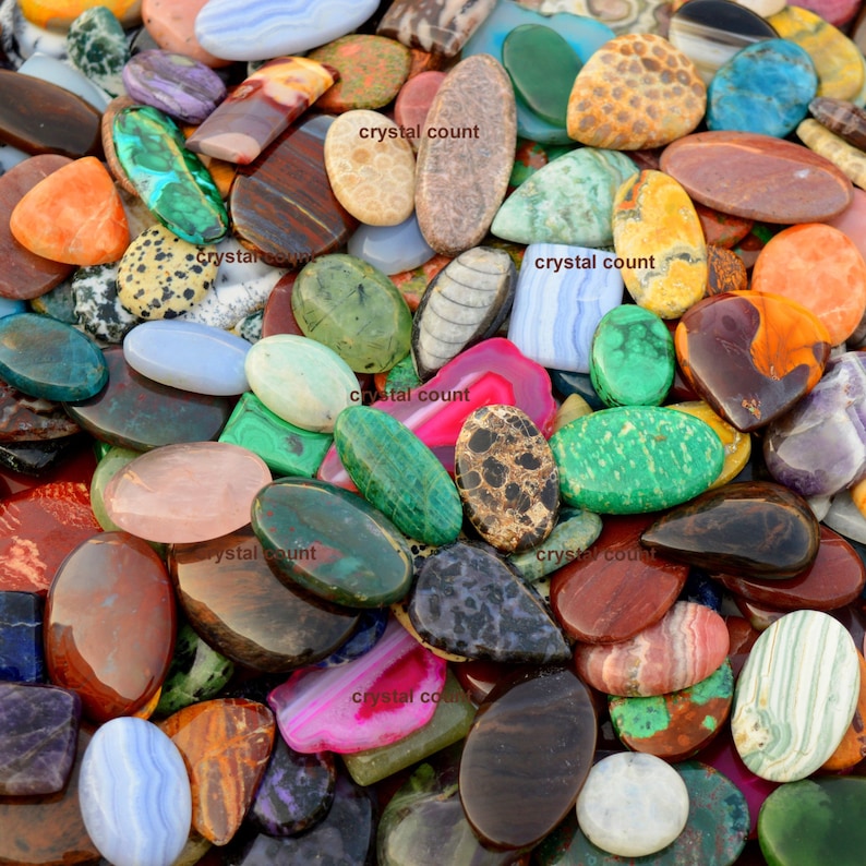 May include: A large collection of colourful, oval-shaped gemstone cabochons. The cabochons are made of various materials, including agate, jasper, and turquoise. The cabochons are arranged in a random pattern, and some of them have a polished finish.
