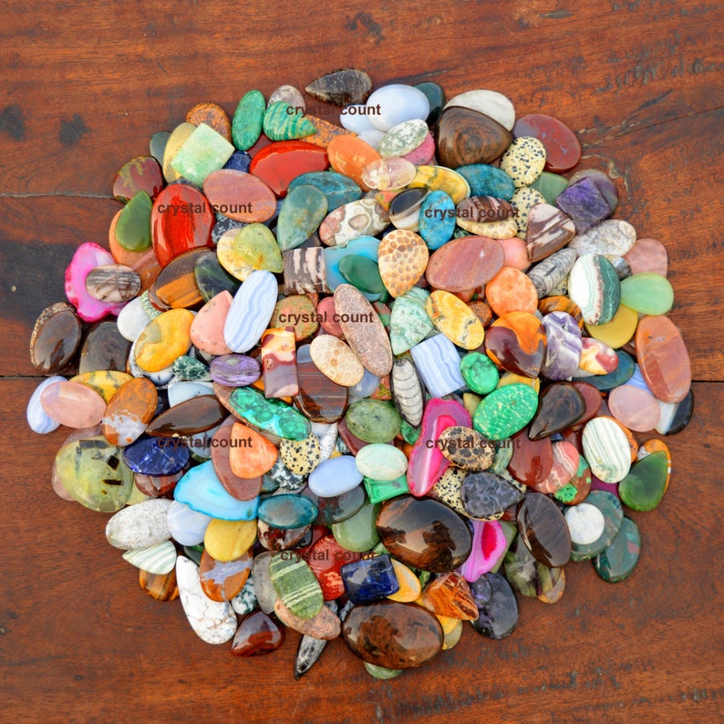 May include: A large collection of colourful, polished, and natural gemstone cabochons. The cabochons are various shapes and sizes, including oval, round, and teardrop. The cabochons are arranged in a circular pattern on a wooden surface.