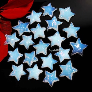 Puede incluir: Una colección de 18 cabujones de ópalo azul iridiscente en forma de estrella. Los cabujones están dispuestos en un patrón aleatorio sobre un fondo negro.