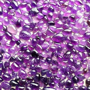 May include: A collection of polished amethyst gemstones in various shapes and sizes, ranging from deep purple to translucent lavender. The gemstones are scattered, creating a textured surface. The text "CrystalCount" is visible.