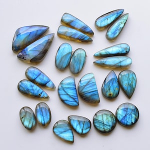 Puede incluir: Una colección de piedras preciosas de labradorita pulida en varias formas, incluyendo gotas, óvalos y semicírculos. Cada piedra muestra una iridiscencia azul vibrante con fracturas internas visibles y tonos base de gris oscuro a marrón.