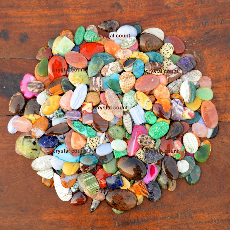 May include: A large collection of colourful, polished, oval-shaped gemstones. The stones are arranged in a circular pattern on a wooden surface. The stones are various shades of red, green, blue, brown, yellow, and orange.