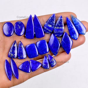 May include: A collection of polished lapis lazuli gemstones in various shapes, including ovals, triangles, and teardrops. The deep blue stones have subtle gold flecks and are displayed against a neutral background.