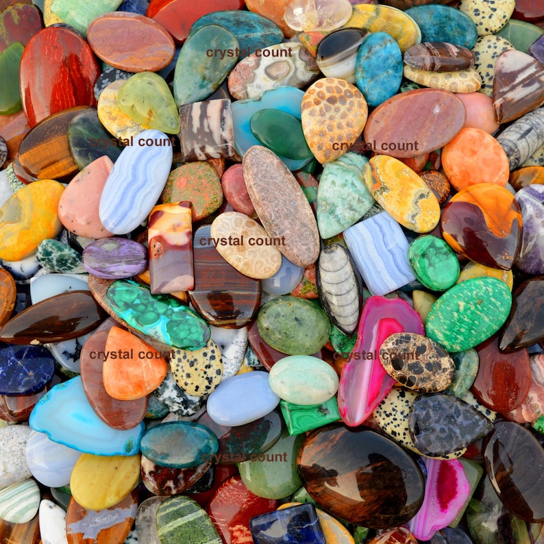 May include: A large collection of colourful, polished, and natural gemstone cabochons. The cabochons are various shapes and sizes, including oval, round, and rectangular. The cabochons are arranged in a random pattern, creating a visually appealing display.
