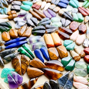 May include: A large collection of assorted gemstone pendants in various shapes and colors, including tiger eye, lapis lazuli, malachite, amethyst, and quartz. Each piece features a small drilled hole for stringing, showcasing natural stone textures.
