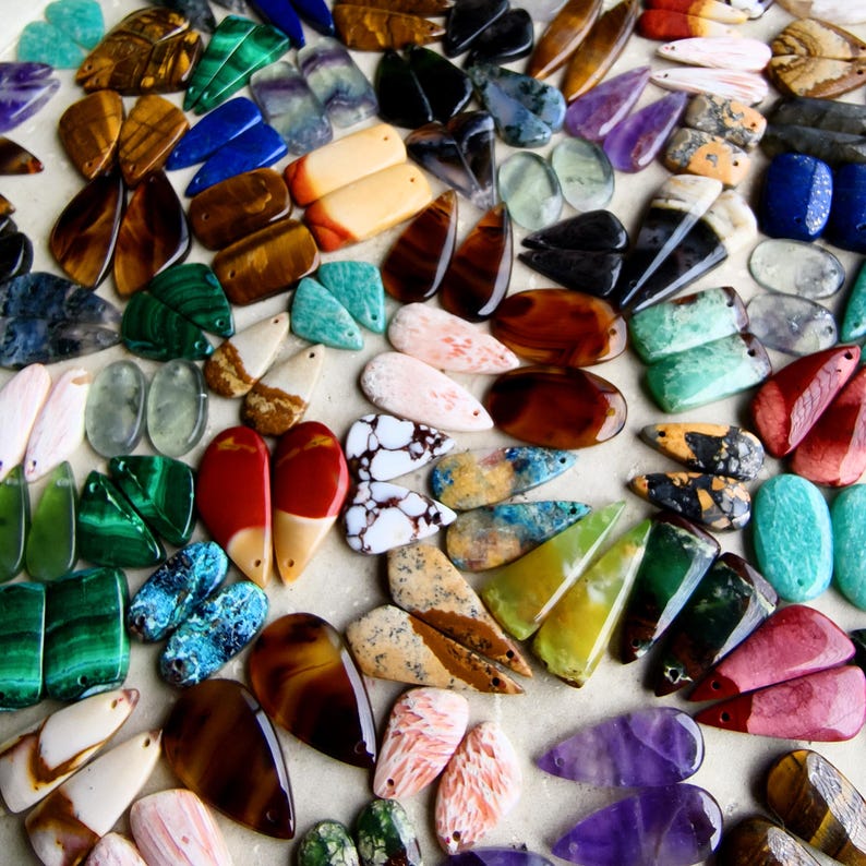 May include: A large collection of polished gemstone cabochons in various shapes, including teardrops, ovals, and rectangles. The stones feature diverse colors and patterns, such as tiger's eye, malachite, amethyst, jasper, and agate, arranged together.