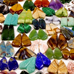 May include: A collection of polished gemstone cabochons in various shapes and colors, including green malachite, blue lapis lazuli, brown tiger eye, turquoise, and white quartz, arranged in pairs on a white surface for jewelry making.