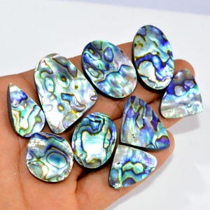 May include: A collection of iridescent abalone shell cabochons in various shapes, including oval, teardrop, and triangular. The shells display a mix of blue, green, and purple hues with a pearlescent sheen. These polished gemstones are ideal for jewelry making.
