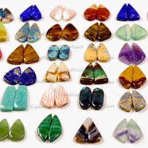 May include: A collection of polished gemstone cabochons arranged in pairs, featuring various shapes and colors including blue, green, brown, purple, and white. Each pair displays unique natural patterns, textures, and mineral inclusions.