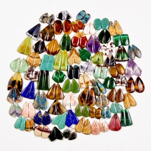 May include: A large collection of polished gemstone cabochons arranged in pairs, featuring a wide variety of colors including green, blue, brown, purple, and white. The stones are shaped as teardrops, ovals, and triangles with smooth surfaces.