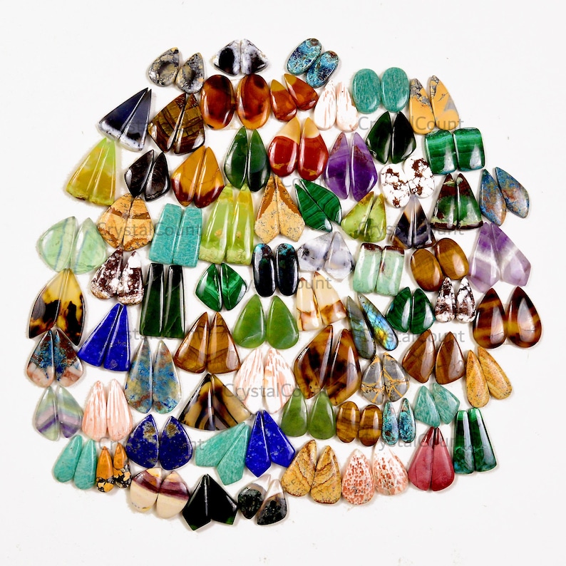 May include: A large collection of polished gemstone cabochons arranged in pairs, featuring a wide variety of colors including green, blue, brown, purple, and white. The stones are shaped as teardrops, ovals, and triangles with smooth surfaces.