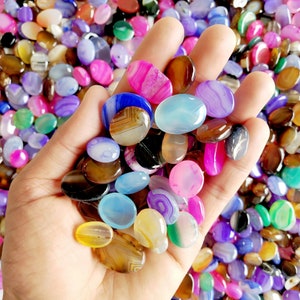 May include: A hand holding a variety of colorful agate stones. The stones are oval shaped and have different patterns and colors, including pink, blue, green, brown, and white.