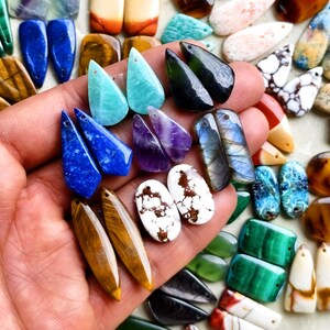 May include: A collection of polished gemstone pendants in various shapes and colors, including blue lapis lazuli, purple amethyst, iridescent labradorite, brown tiger's eye, and white speckled stone, each featuring a small hole for suspension.