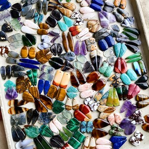 May include: A large collection of assorted gemstone pendants in various shapes, sizes, and colors, including tiger eye, amethyst, malachite, lapis lazuli, and agate, arranged on a flat surface for jewelry making and crafting projects.