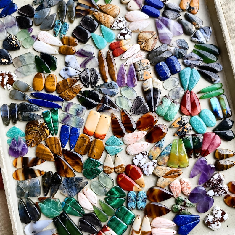 May include: A large collection of assorted gemstone pendants in various shapes, sizes, and colors, including tiger eye, amethyst, malachite, lapis lazuli, and agate, arranged on a flat surface for jewelry making and crafting projects.