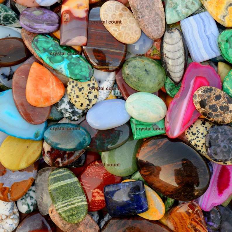 May include: A collection of colourful, polished, and smooth gemstone cabochons. The cabochons are various shapes and sizes, including oval, round, and rectangular. The colours include green, blue, red, orange, yellow, brown, and black.