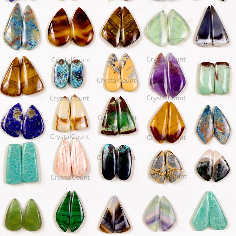 May include: A collection of polished gemstone cabochons arranged in a grid, featuring various shapes including teardrops, ovals, and rectangles. The stones display diverse colors and patterns, including blue, brown, green, purple, and white.