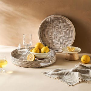 May include: A white wicker tray with a woven pattern, filled with lemons and two clear glass tumblers. A second, larger white wicker tray is in the background, along with a small wooden pedestal with a white plate and a lemon on top.
