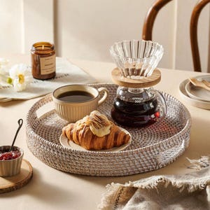 May include: White wicker tray with a coffee pot, a cup of coffee, a croissant, and a candle. The tray is on a white table.