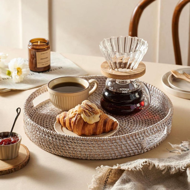 May include: White wicker tray with a coffee pot, a cup of coffee, a croissant, and a candle. The tray is on a white table.
