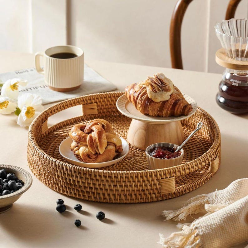 May include: A round woven wicker tray with two handles, featuring a pastry, a small bowl of jam, and a small plate with a croissant on a wooden stand.